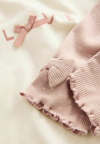 Licht crèmekleurige stof met roze tekst "LOVE" en een strikaccent. Roze geribbelde mouw met een gerimpelde zoom en strikdetail bij de manchette.