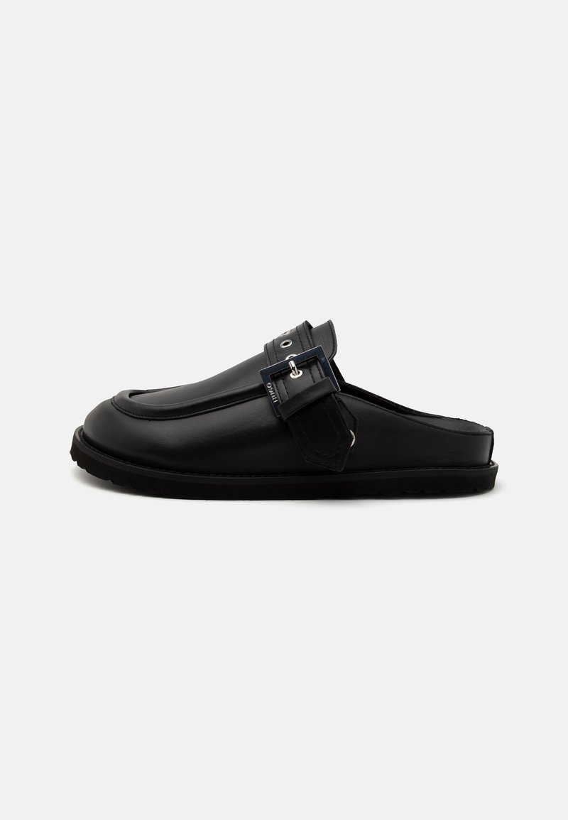 Chaussures noires à enfiler en cuir lisse, avec une sangle avant ornée d'une boucle métallique et une semelle texturée pour une meilleure adhérence.
