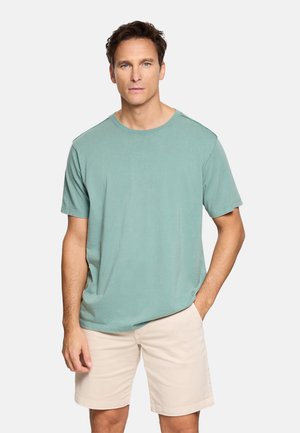 Hombre de pie vistiendo una camiseta de manga corta verde claro y pantalones cortos beige, con una mano en el bolsillo frente a un fondo liso.