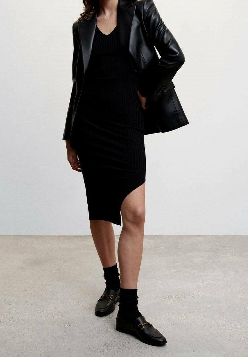 Femme portant une robe noire en maille asymétrique, un blazer en cuir noir, des chaussettes noires et des mocassins noirs, debout sur un sol gris.