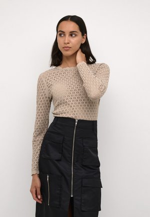 Beige Langarmoberteil mit einem strukturierten, dreieckigen Muster, kombiniert mit einem marineblauen Rock, der einen zentralen Reißverschluss und mehrere Taschen aufweist.