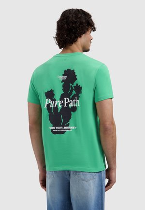 Man met donker krullend haar die een groen T-shirt draagt met een zwarte cactusafbeelding en de tekst "Pure Path" op de achterkant, gecombineerd met lichtblauwe jeans.