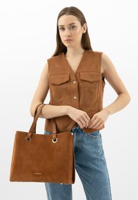 Brązowa torba typu tote z zamszu, z dwoma górnymi uchwytami, z złotymi akcentami i bocznymi detalami; w połączeniu z bezrękawną koszulą i dżinsami.