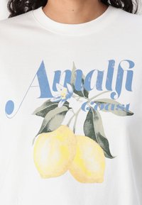 Hvid t-shirt med et grafisk design, der viser to gule citroner og blå tekst, der står "Amalfi Coast", akkompagneret af grønne blade.