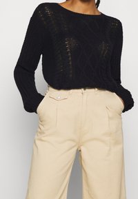 Pull en maille bleu marine foncé avec motifs torsadés et détails en points ouverts, associé à un pantalon beige taille haute avec poches avant et bouton.