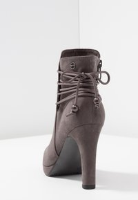 Bottine en suede grise avec un talon haut, présentant des détails lacés, des perles décoratives et une texture lisse, sur fond blanc.