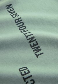 Mintunvihreä kangas, jossa on rohkeat mustat tekstit "TWENTY/FOUR SEVEN" ja "SELECTED." Sileä pinta, jossa hieman kiiltoa.