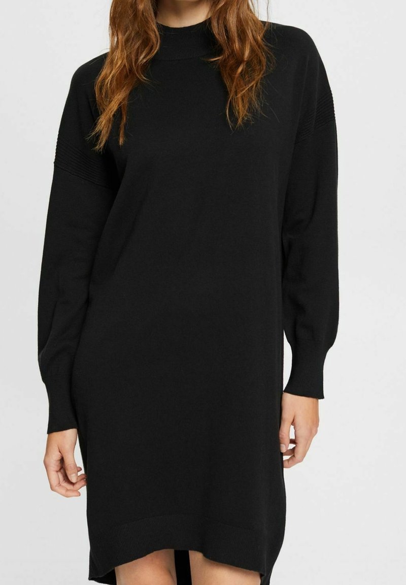 Femme portant une robe-pull noire à manches longues avec des poignets côtelés et un col montant, se tenant devant un fond clair uni.