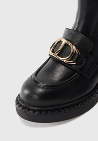 Bottine en cuir noir avec bout rond, présentant un accent de boucle dorée et une semelle texturée avec des détails en relief.