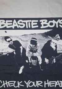 Černobílý grafický tričko s nápisem "BEASTIE BOYS" nad třemi sedícími postavami, s "CHECK YOUR HEAD" níže, vše na šedém pozadí.