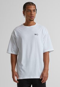 Hvid bomulds T-shirt med korte ærmer, der har et lille sort logo på brystet, afslappet pasform og blød tekstur.