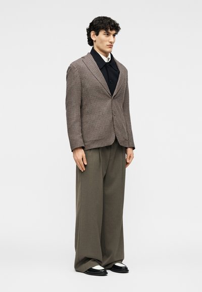 Άνδρας που φοράει ένα καφέ καρό σακάκι πάνω από μαύρο πουκάμισο με κουμπιά, φαρδιά ελιά παντελόνια και δίχρωμα μαύρα και λευκά παπούτσια.