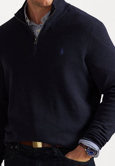 Μπλέ φούτερ με φερμουάρ σε υφασμάτινη υφή με μικρό λογότυπο στο στήθος. Φοράει πάνω από ριγέ πουκάμισο, συνδυασμένο με σκούρο τζιν και καφέ ζώνη.