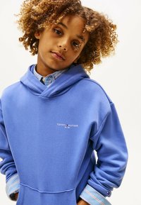 Kind met krullend haar, gekleed in een blauwe Tommy Hilfiger hoodie over een gestreept overhemd, met handen in de zakken, tegen een effen lichte achtergrond.