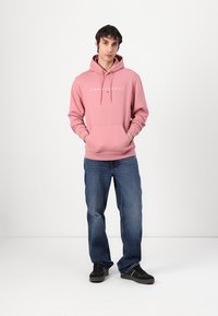 Felpa rosa con tasca frontale e cordino, con il testo bianco "TOMMY JEANS". Abbinata a jeans blu e sneaker nere.