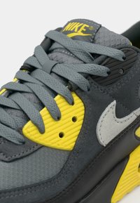 Close-up of a grey and black sneaker featuring yellow accents, textured materials, and prominent Nike branding on the tongue.