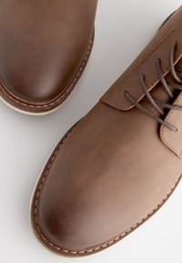 Scarpe in pelle marrone con una texture liscia, punta arrotondata e design con lacci. Cuciture bianche evidenziano i bordi e suola chiara.