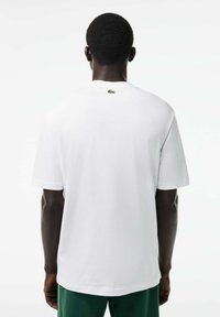Camiseta blanca de manga corta hecha de algodón, con un pequeño logo verde en la parte posterior. Diseño sencillo, corte relajado y textura suave.