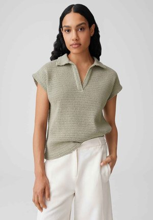 Femme aux longs cheveux bouclés foncés portant un polo à manches courtes en maille vert clair et un pantalon blanc, une main dans la poche.