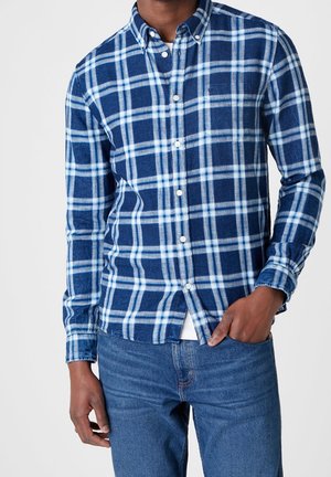 Homme portant une chemise à carreaux bleue et blanche boutonnée sur un sous-vêtement blanc avec un jean bleu, une main dans la poche, sur un fond uni.