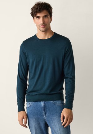 Mand med kort krøllet hår iført en mørk grønblå langærmet sweater og blå jeans, stående mod en ensfarvet lys baggrund.