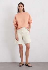 Lichtroze oversized sweatshirt, crèmekleurige shorts en beige sandalen met een gespsluiting. De outfit heeft een ontspannen stijl en zachte texturen.