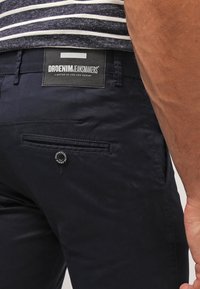 Zbliżenie na osobę w ciemnogranatowych spodniach z tylną kieszenią zapinaną na guzik oraz czarną metką z napisem "DRDENIM JEANSMAKERS."