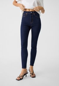 Dark denim high-waisted jeans with a slim fit, featuring contrast stitching and a button closure. Worn with black heeled sandals.