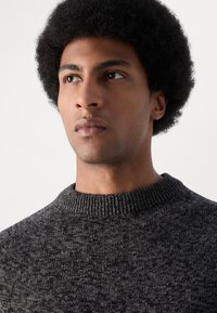 Temno siva pletena sv sweater s rebrastim izrezom, ki ima teksturiran, meliran vzorec. Model ima kratke, kodraste črne lase in nevtralen izraz.