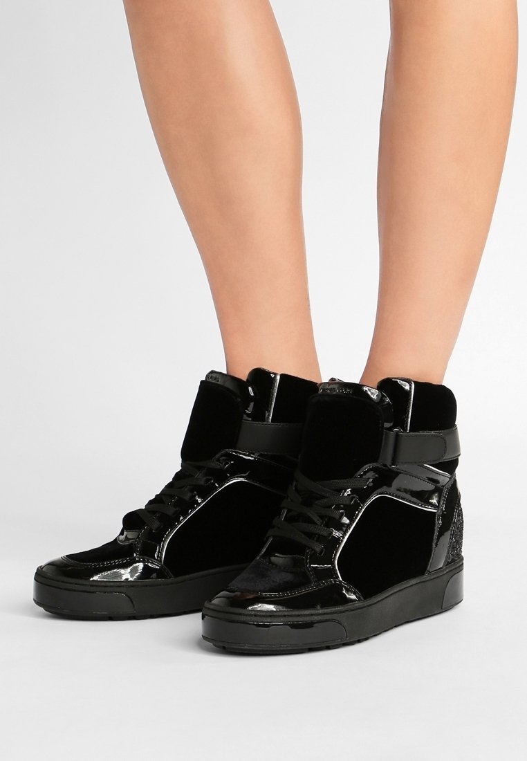 Jambes portant des baskets compensées montantes noires en cuir verni brillant avec des lacets, sur un fond clair uni.