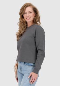 Grijze sweatshirt met een ronde hals en ribgebreide manchetten, gedragen met lichtblauwe jeans. Haar in losse golven gestyled, met een patroonsaccessoire om de taille.