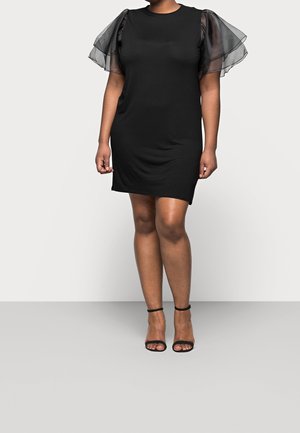 Femme portant une robe noire mi-longue avec des manches bouffantes transparentes et des sandales noires à talons hauts avec bride à la cheville, debout devant un fond uni.