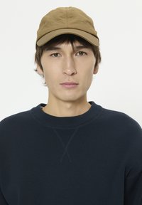 Casquette marron en tissu doux avec un sommet arrondi et une visière courbée, portée sur un sweat-shirt bleu marine à col rond avec un détail de couture prononcé.