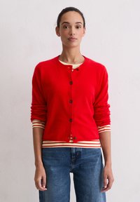 Cardigan en laine rouge avec un col rond et des poignets côtelés, comportant des boutons noirs. Il est superposé à une chemise rayée et associé à un jean bleu.