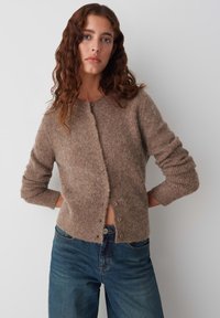 Bruine gebreide cardigan gemaakt van zachte stof, met een ronde halslijn, lange mouwen en knoopsluiting aan de voorkant. Draagt met blauwe jeans.