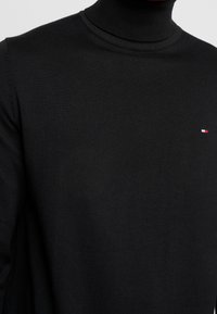 Suéter de cuello alto negro hecho de tela de jersey. Presenta un sutil logotipo rojo, blanco y azul en el lado izquierdo del pecho. Diseño liso y ajustado.