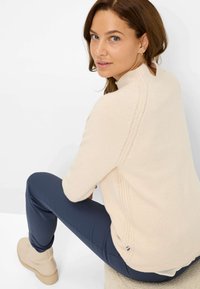 Maglione beige a maglia con scollatura rotonda, cuciture laterali a texture e orlo a costine. Indossato con pantaloni slim-fit blu navy e stivali chiari.