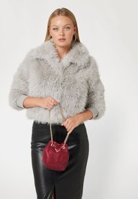 Giacca in pelliccia sintetica grigia con ampio colletto, abbinata a una gonna a tubino in pelle nera, con in mano una borsa a secchiello rossa con paillettes e tracolla in catena.