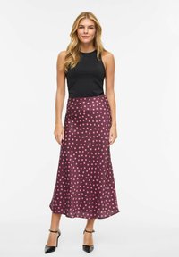 Falda larga de satén en color púrpura oscuro con grandes lunares rosas, combinada con una camiseta sin mangas negra ajustada y zapatos de tacón negros.