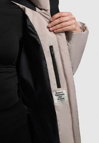 Hosszú, párnázott dzseki bézs külsővel és fekete belsővel, cipzáras zsebbel és egy címkével, amelyen ez áll: "Egy meleg ölelés textilből."