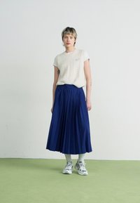 Plissé mørkeblå midi-skirt kombinert med en kremfarget kortermet kabelstrikket topp. Tilbehør inkluderer hvite sokker og grå joggesko.