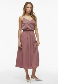 Liliowa, bezrękawna sukienka maxi z dekoltem w kształcie litery V i elastyczną talią; gładki materiał opada do połowy łydki. Noszona z kremowymi czółenkami na szpilce.