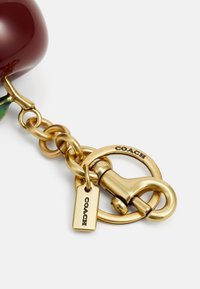 Charme de pomme rouge profond et brillant attaché à une chaîne couleur or avec un clip rond et une étiquette rectangulaire gravée "COACH."