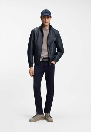 Man met een marineblauwe pet, zwarte leren jas, grijze trui, marineblauwe broek en beige sneakers staat met één hand in zijn zak.