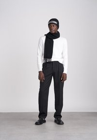 Beanie negru cu logo-ul "Moschino", cămașă albă cu mâneci lungi, eșarfă neagră cu ciucuri, pantaloni negri croiți, și pantofi negri.