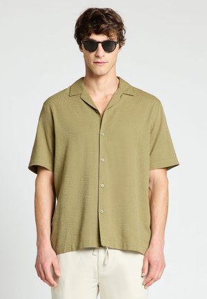 Uomo con occhiali da sole neri, camicia verde oliva a maniche corte con fantasia sottile e pantaloni beige chiaro su sfondo uniforme.