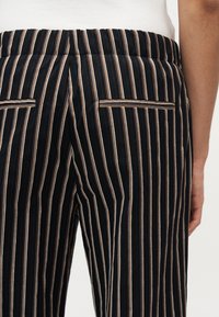 Κοντινό πλάνο μαύρων παντελονιών με κάθετες καφέ και λευκές ρίγες και δύο οριζόντιες πίσω τσέπες, που φοριούνται από άτομο με καστανό χρώμα δέρματος.