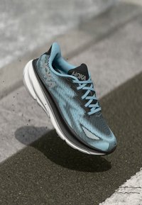 Hoka-sneakers i blått og svart med teksturert strikket overdel, karakteristisk logo og polstret såle for støtte, vinklet mot fortau.