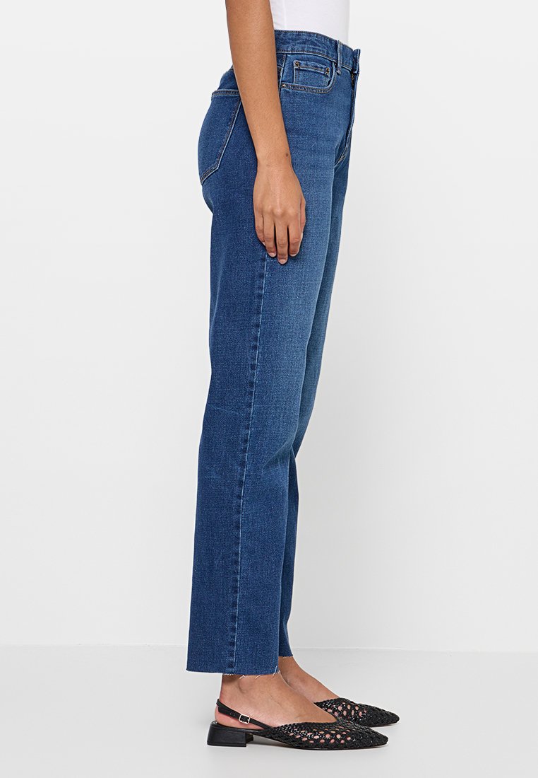 Dunkelblaue gerade geschnittene Jeans mit mittelhohem Bund, die ein klassisches Fünf-Taschen-Design und dezente Nähdetails aufweisen. Getragen mit schwarzen Ballerinas.