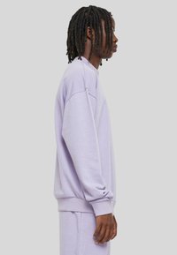 Sweatshirt violet avec une coupe décontractée, des poignets et un col côtelés. Tissu doux et texture lisse, présenté de côté sur un fond neutre.